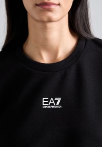 Felpa nera con collo tondo, caratterizzata da un logo bianco EA7 Emporio Armani. Il tessuto appare liscio e senza ornamenti.