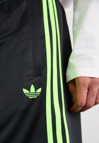 Fekete sportnadrág, amelyen három függőleges neon zöld csík és egy azonos színű logó található a bal combon; az anyag sima és könnyű.