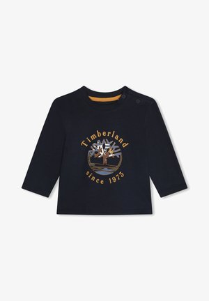 Camisa de manga larga de color azul marino hecha de algodón. Presenta un logotipo circular de Timberland en dorado con una imagen de un árbol, y el texto "desde 1973" abajo.