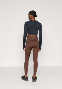 Marineblauw crop top met lange mouwen, bruine high-waisted legging, zwarte sportschoenen. De legging heeft een subtiel logo en een aangesloten pasvorm.