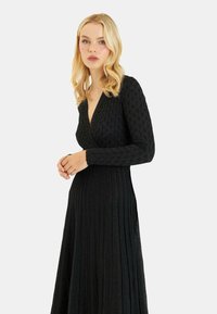 Schwarzes Strickkleid mit tiefem V-Ausschnitt, langen Ärmeln und Faltenrock. Verfügt über ein strukturiertes Muster und eine glatte Oberfläche.