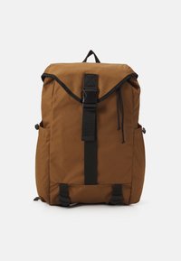 Mochila de tela marrón con un diseño de parte superior enrollable, cierre de hebilla y correa negra, bolsillos laterales y fondo reforzado. Estética minimalista.