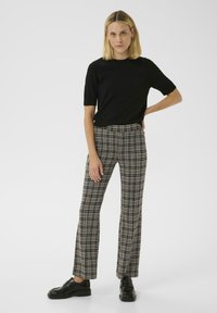 Svart kortärmad topp i kombination med rutiga byxor med ett mönster av beige, svart och vitt. Modellen bär svarta skor. Vit bakgrund.