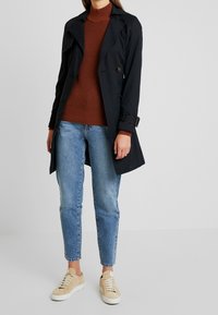 Marineblauwe trenchcoat met dubbelgeknoopte voorkant, bruine geribbelde trui, lichtblauwe spijkerbroek en beige sneakers; voorzien van een ceintuur in de taille en een klassieke kraag.