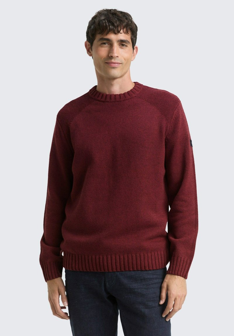 Burgunderfarbener Strickpullover mit rundem Ausschnitt und gerippten Bündchen, mit einem strukturierten Muster an den Schulternähten. Getragen über dunklen Jeans.