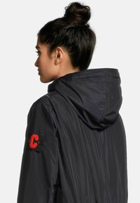 Veste à capuche noire avec une texture lisse et brillante, ornée d'un écusson rouge "C" sur la manche gauche. Le design présente une forme simple et épurée.