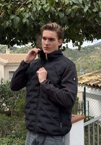 Man die de kraag van een zwart jack met ritssluiting rechttrekt, staand buiten bij groen, een hek en gebouwen met pannendaken.