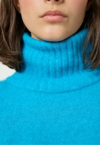 Tirkīza krāsas augsto apkakli svīteri mīkstā, teksturētā adījumā ar ribota apkakli un korpusu, rādot vienmērīgu krāsu un vienmērīgas šuves.