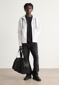 Haut à capuche zippé blanc avec des cordons noirs sur un t-shirt noir, associé à un jean sombre et des baskets noires, tenant un sac de sport noir.