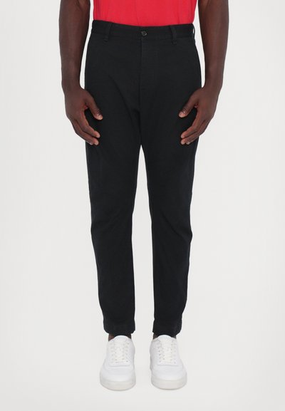 Pantalons noirs cintrés en tissu léger, dotés d'une fermeture à bouton, de poches et de poignets élastiques, associés à des baskets blanches.