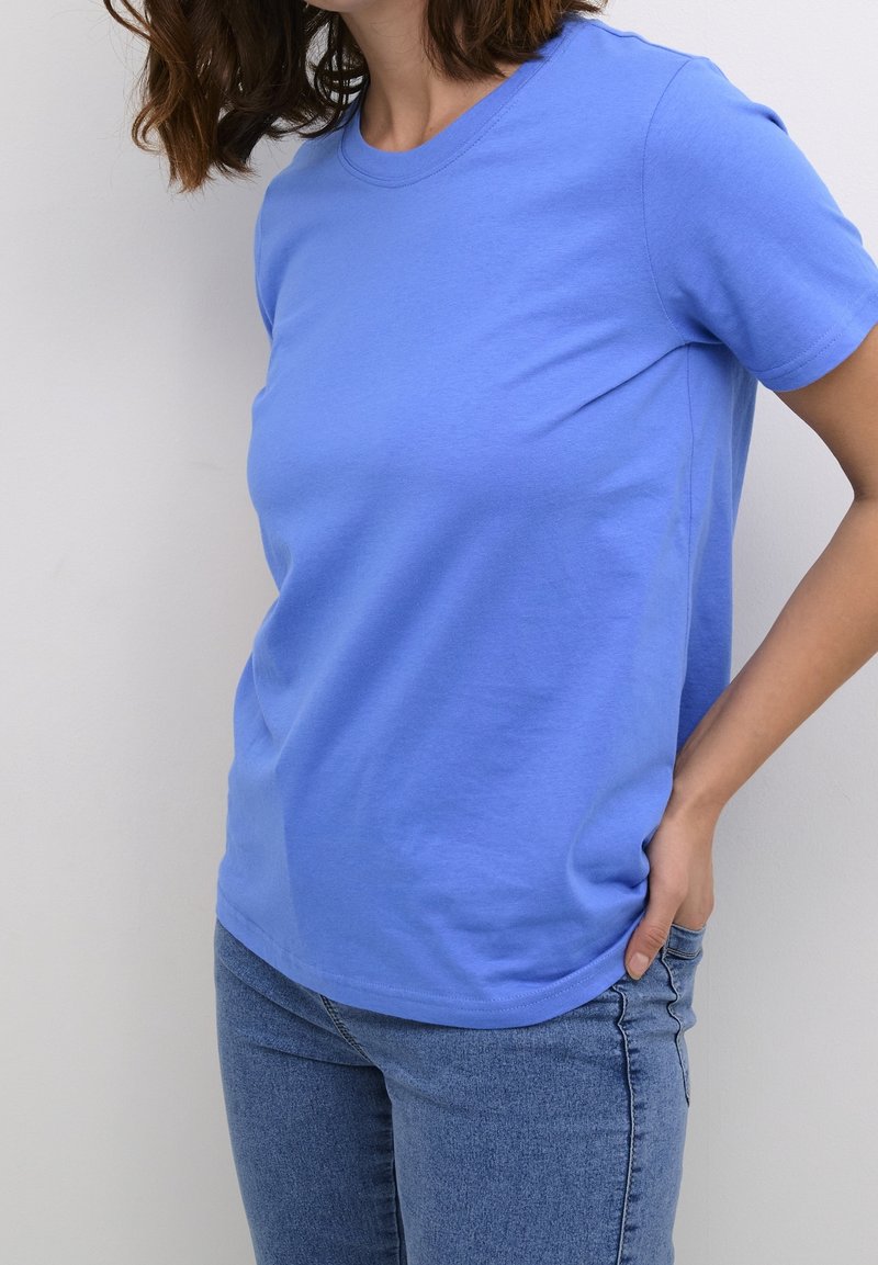 Personne portant un t-shirt bleu uni à manches courtes et un jean bleu, une main dans la poche, visible du cou aux hanches.
