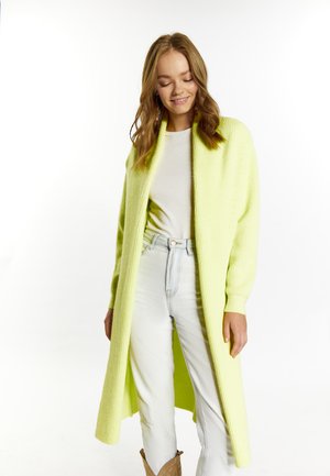 Long gilet vert clair avec texture côtelée et devant ouvert. Porté par-dessus une chemise blanche et un jean bleu clair. Design doux et décontracté.