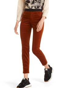 Pantalon en velours côtelé couleur rouille, coupe ajustée, poches avant et taille mi-haute, associé à des baskets noires et un haut à motifs.