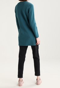 Tyrkysový dlouhý rukáv s texturovaným vzorem, spárovaný s černými slim-fit kalhotami a světle růžovými botami. Halenka má kulatý výstřih a boční kapsy.