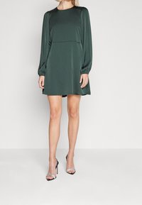 Gaun lengan panjang hijau tua dengan leher bulat, bahu berlipit, dan potongan santai, dipadukan dengan sandal hak tinggi hitam.