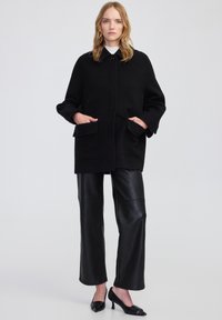 Cappotto di lana nero con colletto, due tasche frontali e una vestibilità comoda. Abbinato a pantaloni neri in pelle con gamba ampia e scarpe con tacco basso.