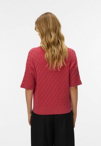 Roza pleten pulover s kratkimi rokavi, teksturiranim diamantnim vzorcem, sproščenim krojem in rebrastim robom. Nošen s črnimi širokimi hlačami.