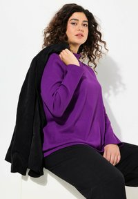 Lila Langarmoberteil mit Stehkragen, kombiniert mit einer schwarzen Cordjacke, die über eine Schulter drapiert ist, und schwarzen Hosen. Glatte Textur.