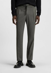 Gray tailored trousers with pleats, made from textured fabric, featuring an elastic waistband and drawstring. Black dress shoes complement the look.
