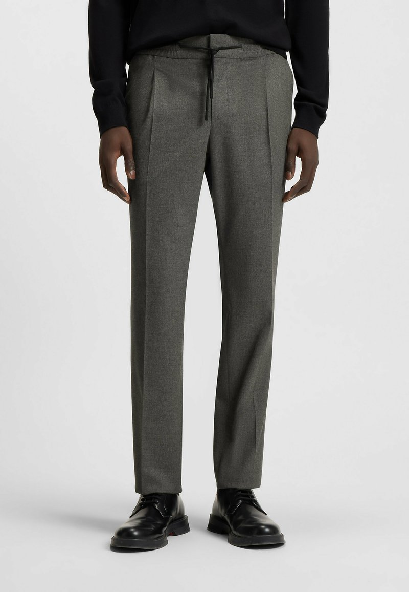 Gray tailored trousers with pleats, made from textured fabric, featuring an elastic waistband and drawstring. Black dress shoes complement the look.