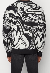 Pull à motif marbré noir et blanc porté par une personne de dos, associé à un jean noir.
