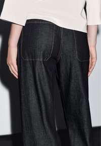 Černé džíny s zadními kapsami, kontrastním prošitím a volným střihem. Tkanina má hladkou strukturu a jemný lesk.
