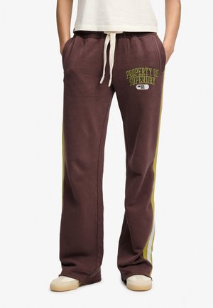 Osoba ubrana w brązowe dresowe spodnie z żółtymi paskami po bokach, białym ściągaczem, napis "Property of Superdry" na lewej udzie, białe trampki i biały top.