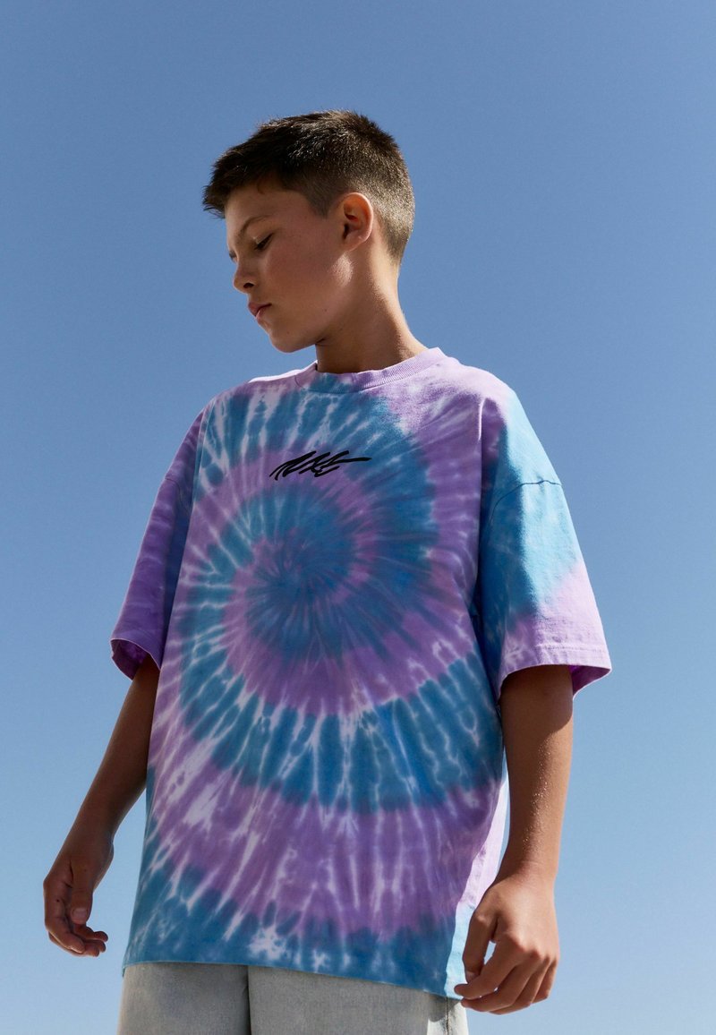 Värviline tie-dye t-särk liljade ja siniste toonidega, millel on spiraalne muster. Lühikesed varrukad, mugav lõige, rinnal väike must graafika.
