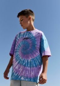 Tricou tie-dye în nuanțe de mov și albastru cu un model în spirală. Mâneci scurte, croială relaxată, având o mică grafică neagră pe piept.