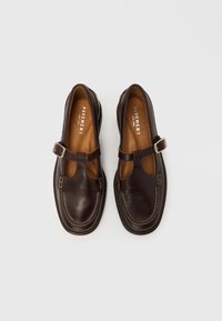 Chaussures en cuir marron de style Mary Jane, avec une bride supérieure munie d'une boucle argentée et des coutures blanches le long des bords.