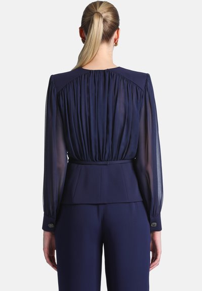 Femme avec queue de cheval blonde portant un chemisier bleu marine à manches longues transparentes et un pantalon assorti, vue de dos.