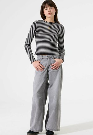 Jonge vrouw staat met handen in de zakken, gekleed in een zwart-wit gestreepte lange mouw top, grijze wijde spijkerbroek en zwarte schoenen.