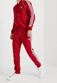 Punainen urheiluasu, joka on valmistettu teksturoidusta kankaasta ja sisältää valkoiset kolme viivaa hihassa ja housuissa. Mukana on vetoketjullinen takki ja elastinen vyötärö.