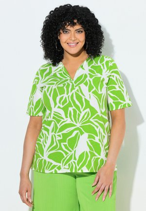 Lächelnde Frau mit lockigem schwarzen Haar, die Creolen trägt, ein grün-weißes kurzärmeliges Hemd mit Blattmuster und hellgrüne Hose.