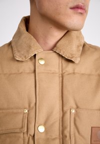 Braune gesteppte Jacke mit einem Cordkragen, goldenen Druckknöpfen und frontalen Taschen mit einem Lederlogo-Patch.