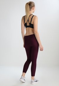 Top sportivo nero con bretelle incrociate sul retro e leggings color marsala. I leggings sono attillati e lisci, abbinati a scarpe da ginnastica grigie.