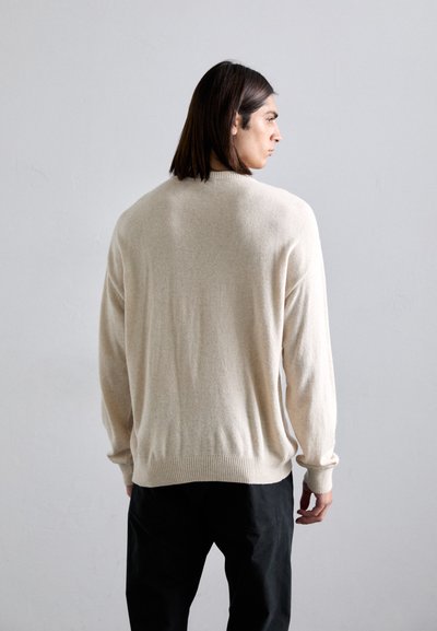 Beige stickad tröja med långa ärmar, rundad halsringning och ribbad nederkant. Mjuk textur och avslappnad passform. Bärs med svarta byxor.