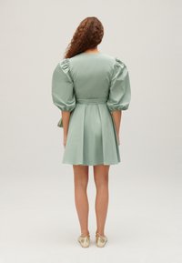Mátově zelené šaty s nabíranými rukávy, přiléhavým pasem a rozšířenou sukní. Hladká textura látky a délka ke kolenům. Zlaté boty s mašlemi.