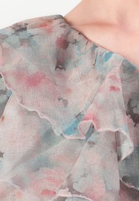 Transparent mångfaldigt lager tyg med volanger i mjuka pastellblommönster, som täcker axeln och nyckelbenet.