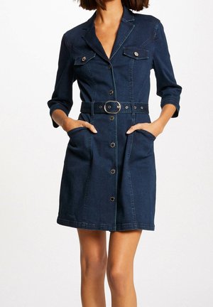 Kvinde iført en mørkeblå denimkjole med knapper og trekvartærmer, bælte med rund spænde, to brystlommer, og hænderne i forlommerne.