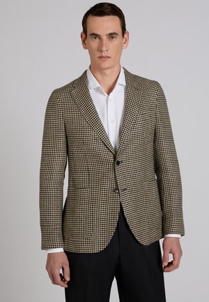 Mężczyzna w beżowo-czarnej marynarce w pepitkę, białej koszuli i czarnych spodniach, stojący na tle szarego, jednolitego tła.