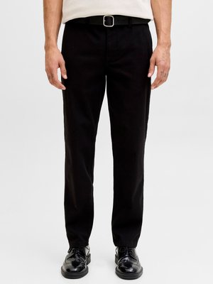 Homme portant un pantalon de costume noir, une ceinture noire et des chaussures de ville noires et brillantes, debout devant un fond blanc uni.