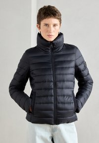 Melnā pūkaina jaka ar augstu apkakli, kvadrātveida dizainu, priekšējo rāvējslēdzēju un sānu kabatām. Materiāls izskatās viegls un gluds.