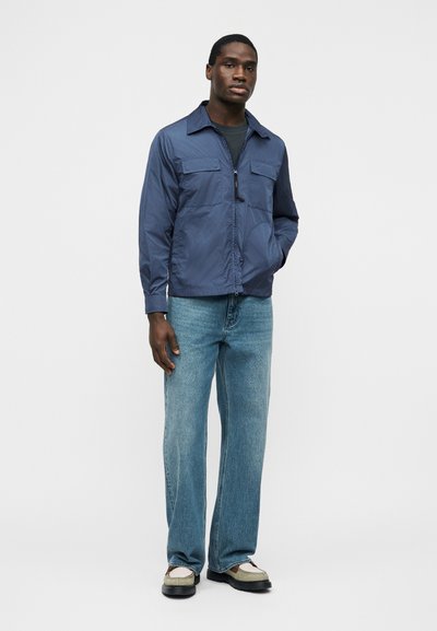 Homme portant une veste zippée bleue, un t-shirt noir, un jean bleu clair et des chaussures beiges, debout avec une main dans la poche sur fond blanc.
