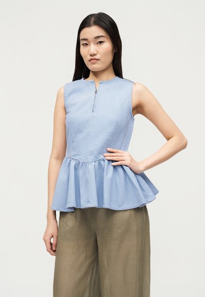Jeune femme aux longs cheveux foncés pose en portant un top peplum sans manches bleu clair et un pantalon large beige ample.