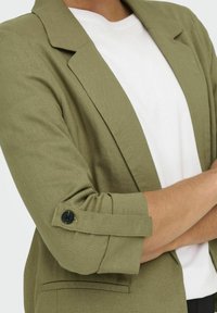 Blazer verde oliva con un design strutturato, polsini a due bottoni e una t-shirt bianca sotto. Tessuto strutturato e vestibilità su misura.