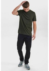 T-shirt corto verde scuro, taglio classico, abbinato a pantaloni neri e sneakers grigio-blu, in piedi con una mano sulla testa.