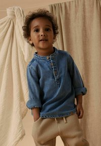 Gaiši zilas krāsas džinsa krekls ar garām piedurknēm, pogu aizdari un uzvītām piedurknēm, kopā ar bēšas krāsas teksturētām jogger biksēm. Fonā redzami mīksti audumi.