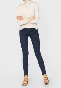 Top de cuello alto beige, diseño ajustado, combinado con jeans ajustados azul marino oscuro y sandalias marrones con tiras y detalles dorados.