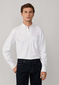 Camisa blanca de botones con un bolsillo en el pecho, hecha de algodón. Presenta un cuello clásico y mangas largas. Combinada con jeans de mezclilla oscura.